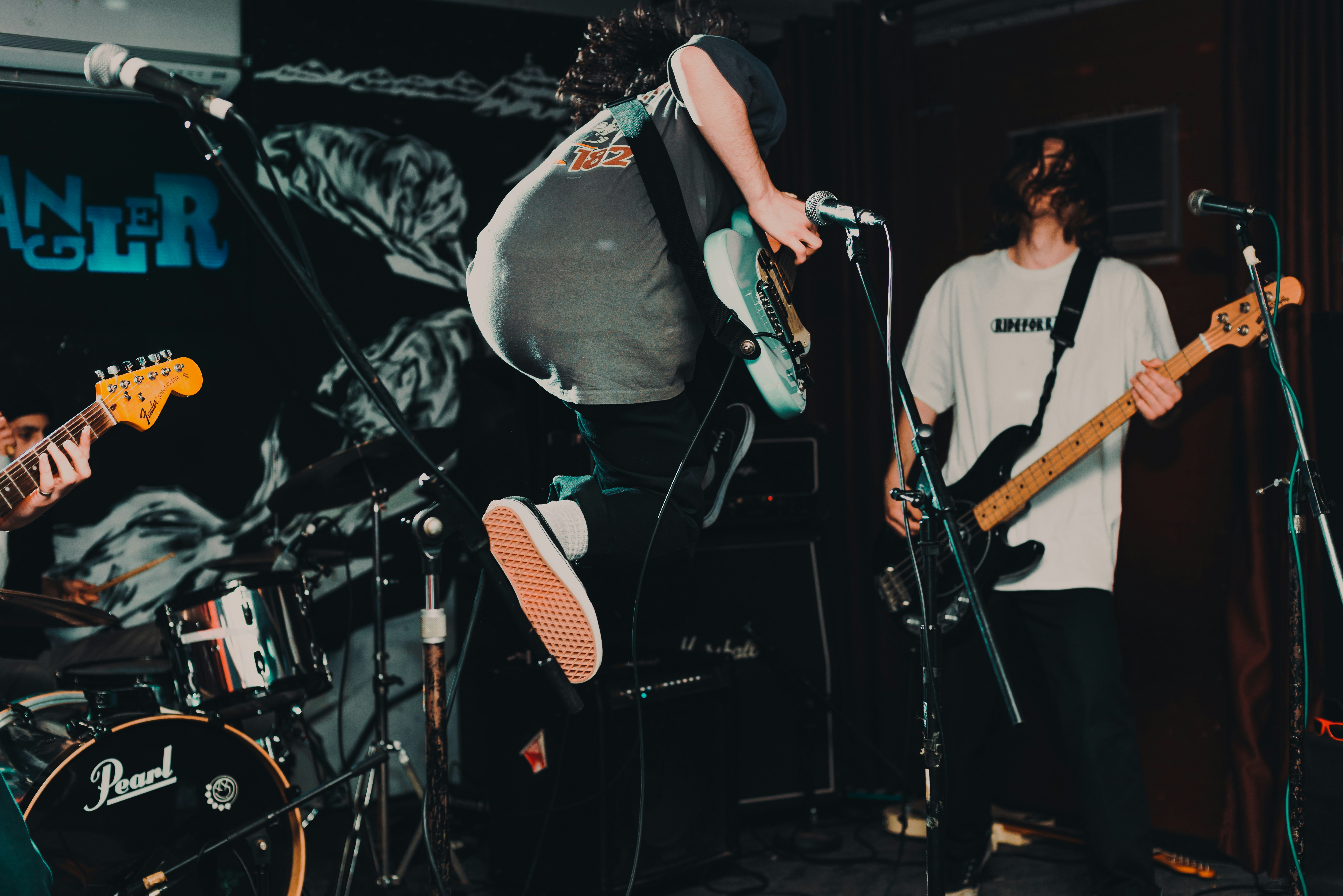 The image shows a Metal band performing onstage, three guitarists are in view with the middle guitarist jumping in the air while playing guitar.