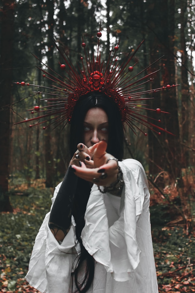 Woman standing in the woods, with a red pointed headpiece, long black hair and a white gown, holding her hands in front of her. Her eyes are white with no iris or pupil