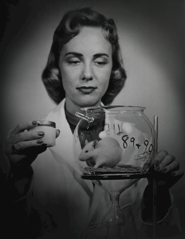 1950s styled woman holding glass bowl with a mouse in it. Likely in an experitmental laboratory.