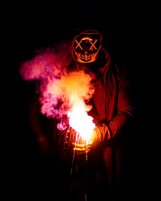 Person in hoodie holding fire and smoke, wearing a black leather mask with white X's drawn on the eyes. Chilling picture